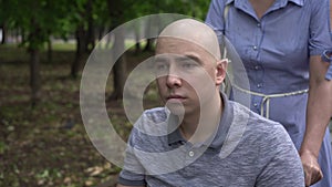 A woman is driving a young man with oncology in a wheelchair through the park closeup. The man is bald due to