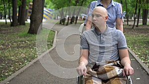A woman is driving a young man with oncology in a wheelchair through the park. The man is bald due to chemotherapy.