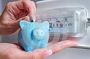 A woman drops a coin into a blue piggy bank near a natural gas meter.