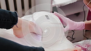 Woman is drying shellac on toes in uv lamp in beauty salon, closeup view. Pedicurist master is covering painting client