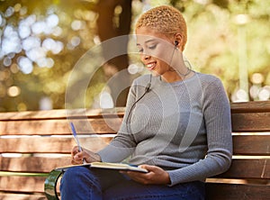 Woman, earphone and writing in book, diary or journal in a park outdoor with a happy smile on a bench. Latino girl write