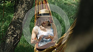 Woman in earphones lying in hammock and listening to music at green garden. Brunette female listens to music from earphones, using