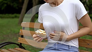 Woman eating burger in park, feeling nausea, food poisoning symptom, gastritis