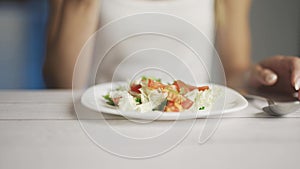 A woman is eating fresh vegetable salad while sitting in her kitchen. Slim woman is observing diet and counting calories