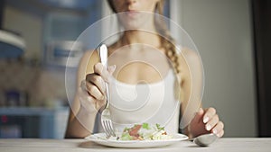 A woman is eating fresh vegetable salad while sitting in her kitchen. Slim woman is observing diet and counting calories