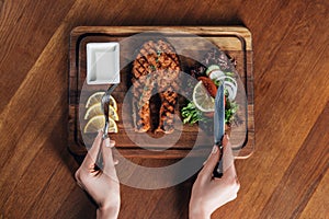 Woman eating grilled salmon steak served on wooden board with lemon and lettuce