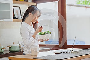 Woman eating healthy salad in kitchen with laptop