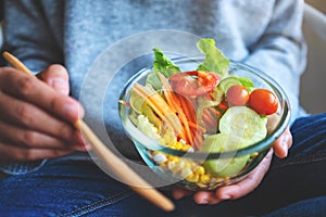 A woman eating and holding a bowl of fresh mixed vegetables salad by fork