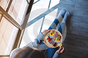 A woman eating and holding a wooden plate of fresh mixed fruits on skewers while sitting on the floor