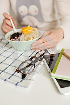 Woman eating porridge and fruits, notepad tablet and glasses on