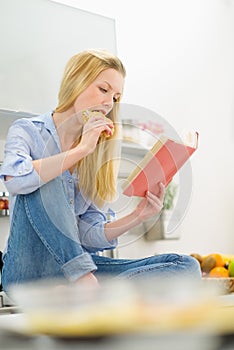 Woman eating sandwich and reading book in kitchen