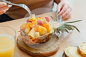 Woman eating tasty tropical fruit salad in half pineapple bowl. Berries raspberries pineapple tangerines oranges in