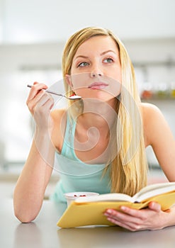 Woman eating yogurt in kitchen and reading book