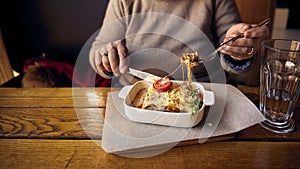 A woman eats lasagnia with cheese and a Behamel sauce from a white ceramic plate located next to a glass of water on the table