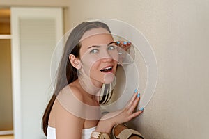 Woman eavesdropping through the wall of the room through a glass cup, curiosity, espionage, surprised face, vivid emotions