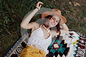 A woman in eco-friendly clothing in a hippie look is lying on a colored plaid smiling and looking at an autumn sunset in