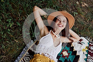 A woman in eco-friendly clothing in a hippie look is lying on a colored plaid smiling and looking at an autumn sunset in