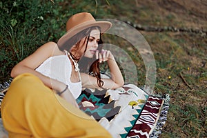 A woman in eco-friendly clothing in a hippie look is lying on a colored plaid smiling and looking at an autumn sunset in