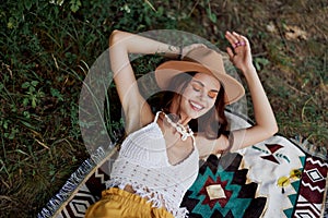 A woman in eco-friendly clothing in a hippie look is lying on a colored plaid smiling and looking at an autumn sunset in