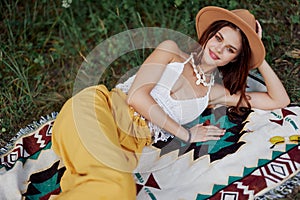 A woman in eco-friendly clothing in a hippie look is lying on a colored plaid smiling and looking at an autumn sunset in