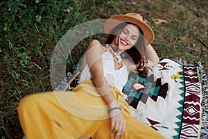 A woman in eco-friendly clothing in a hippie look is lying on a colored plaid smiling and looking at an autumn sunset in