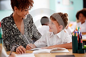 Woman Elementary School Teacher Giving Male Pupil Wearing Uniform One To One Support In Classroom