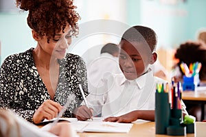 Woman Elementary School Teacher Giving Male Pupil Wearing Uniform One To One Support In Classroom