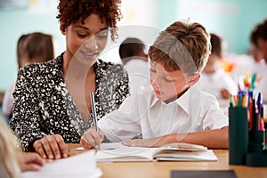 Woman Elementary School Teacher Giving Male Pupil Wearing Uniform One To One Support In Classroom