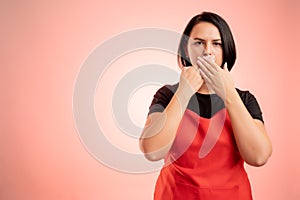 Woman employed at supermarket with red apron and black t-shirt covering mouth like mute concept