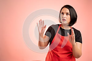 Woman employed at supermarket with red apron and black t-shirt showing fear, with her arms up