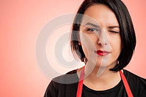 Woman employed at supermarket with red apron and black t-shirt winking one eye