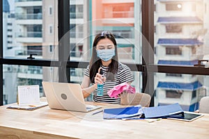 Woman employee wearing face mask preparing alcohol spray and cloth for cleaning on wooden desk in the office