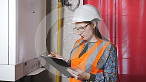 A woman engineer checks the parameters of the heating system. The engineer in the helmet writes down instrument readings