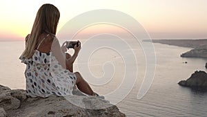 Woman enjoy the view of the sea during sunset time and taking photo on cellphone. Longhaired girl sitting on the cliff