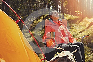 Woman enjoying the nature tranquility while resting in camping chair with hot drink at forest camp. outdoor adventure