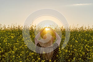 Woman enjoying spring making a heart with hands against sunset sun light in yellow flowers field.Spring love concept , copy space