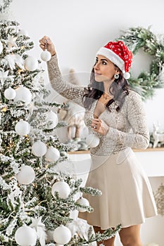 Woman enjoys Christmas preparation wearing Santa& x27;s hat putting decorations on an x-mas tree