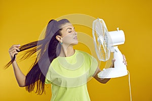 Woman enjoys fresh air and escapes summer heat with electric fan isolated on orange background.
