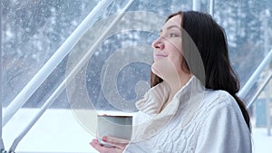 Woman enjoys hot drink looking at winter in panoramic window