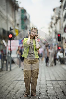 A woman enjoys music on headphones while standing on a busy street.