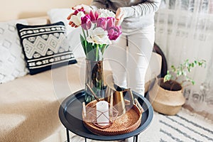 Woman enjoys tulips flowers put in vase on table with tray, watering can and candle. Interior and spring decor at home