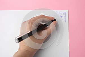 Woman erasing web drawn with erasable pen on sheet of paper against pink background, top view