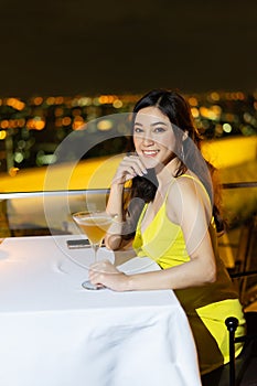 Woman in evening dress holding cocktail at restaurant over night city background