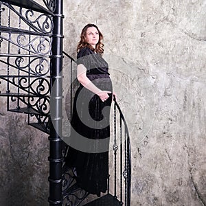A woman in an evening dress on the steps of a black spiral staircase, vintage full-length portrait of a pregnant woman