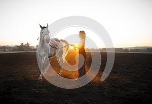 Woman in evening dress working with grey Arabian horse on a black burnt field backlit with setting sun