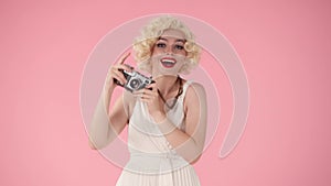 Woman excitedly taking pictures on old camera, says wow. Woman looking like Marilyn Monroe in studio on pink background