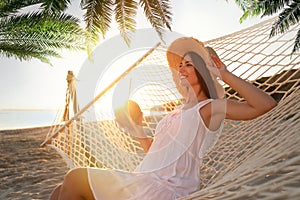 Woman with exotic cocktain relaxing in hammock under green palm leaves on sunlit beach