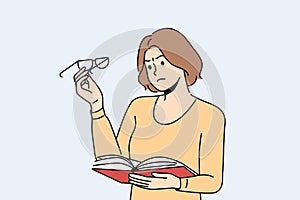 Woman experiences vision problems reading book and looks at glasses with upset grimace