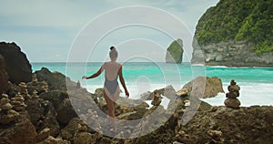 Woman exploring pristine beach by ocean cliffs. Adventurer girl in swimsuit walking by stone stacks and sea spray.