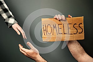 A woman extends a helping hand to a man holding a cardboard box with the text homeless. Dark background of a greenish